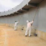 Aplicação do sistema fiberglass em parede de concreto por trabalhadores utilizando rolos com cabo extensor em estrutura industrial.